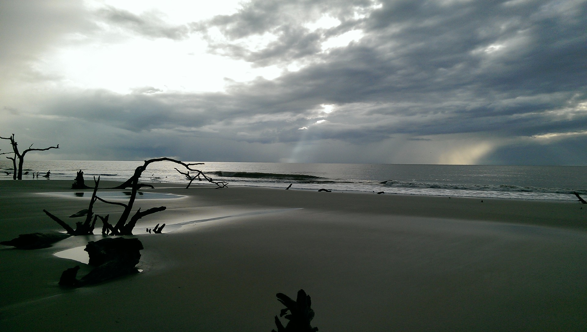 Archival St. Catherines Island shoreline with driftwood, wet sand and surf beneath passing storm clouds Photograph: William D Bone · CC BY-SA 3.0. Historical view; current conditions may differ.
